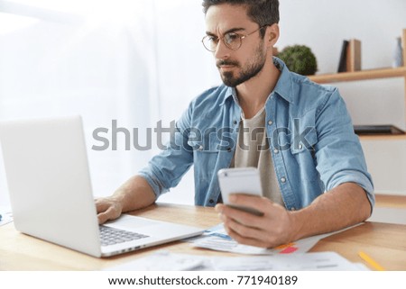Young serious handsome male enterpreneur being busy as works with statistics data and analyzes perfomance, types on laptop computer and messages with partners on smart phone. Business, communication