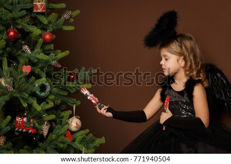 x-mas, winter vacation and people concept - little girl in black angel costume sits on a trunk near a Christmas tree and looks at a toy