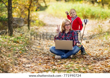 A Mom and  boy take pictures of themselves on  smartphone, smiling happily at  camera. Mom holds a laptop on his lap, and his son hugged her from behind, background  very fuzzy autumnal