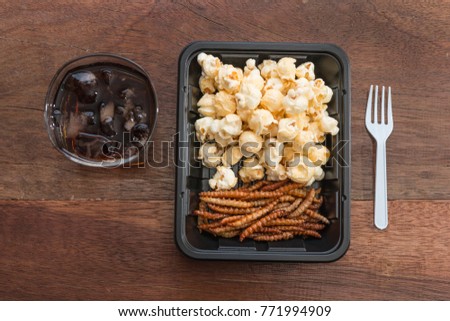 Popcorn with bamboo worm fried insect on the black plate with fresh cola drink on wooden table background. Insect food is the healthy meal high protein diet concept. Top view.
