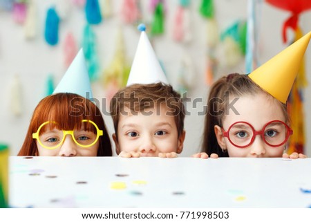 Two adorable girls in eyewear and one boy play game together, celebrate festive event, hide behind table, have cheerful funny expressions, look directly into camera. Childhood and celebration