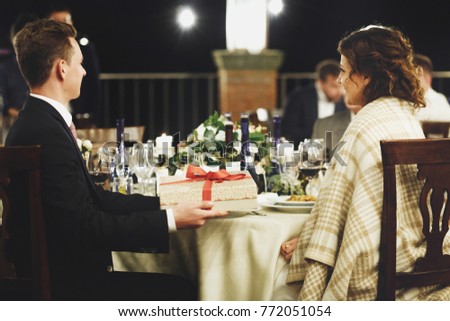 Groom holds a present box before a bride sitting at dinner table