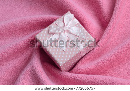 A small gift box in pink with a small bow lies on a blanket of soft and furry light pink fleece fabric with a lot of relief folds. Packing for a gift to your lovely girlfriend