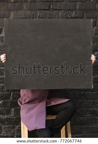 background, advertisment, equipment concept. woman wearing lilac autumn coat is holding not big blackboard without any sign on it and hiding behind it, she is sitting on the chair