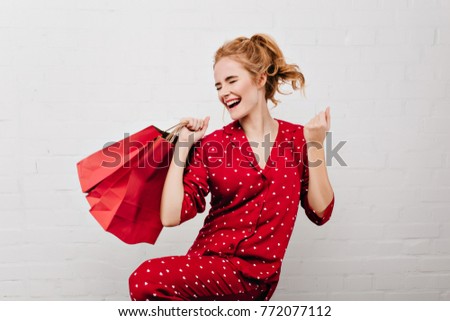Enthusiastic girl in red pajamas dancing with paper bags isolated on white background. Funny blonde woman holding new year gifts, standing near brick wall.