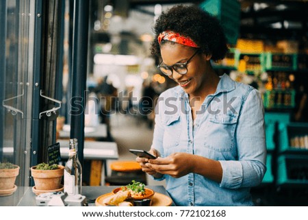 Stylish young African woman smiling while sitting alone at a counter in a bistro taking photos of her food with her smartphone