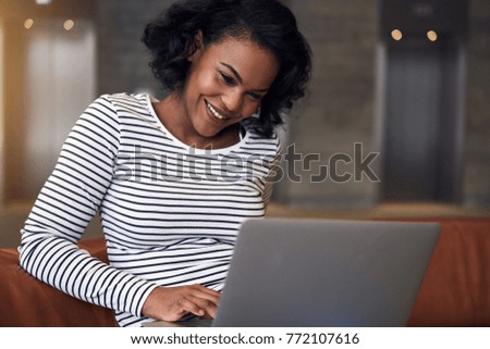 Smiling young African college student sitting alone on a sofa on campus studying online with a laptop