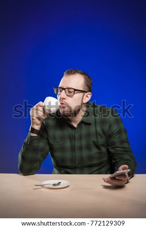 Emotional man in glasses is drinking coffee from a white cup at the table and posing on a blue background