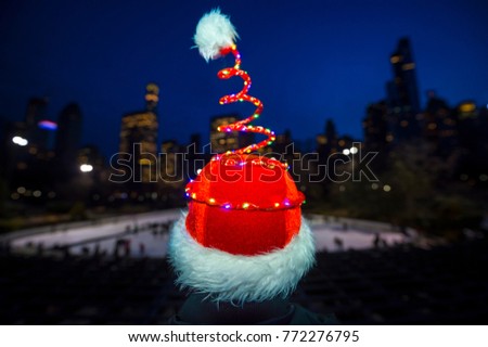 Colorful defocused holiday lights glow on a springy Santa hat in front of a twinkling city skyline and ice rink background