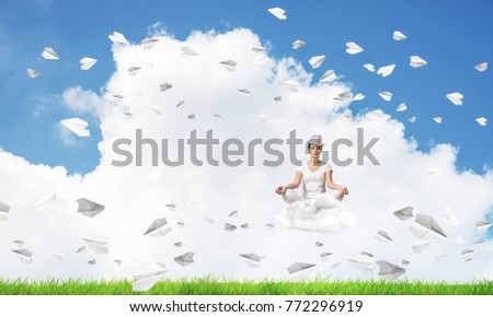 Young woman keeping eyes closed and looking concentrated while meditating on cloud among flying paper planes with bright and beautiful landscape on background.