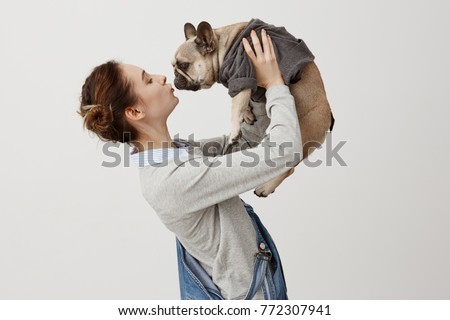 Horizontal shot of adult girl in jeans overalls kissing cute puppy while raising it in air. Young girl being in love with her dog clothed in trendy jumper. Display of affection Royalty-Free Stock Photo #772307941
