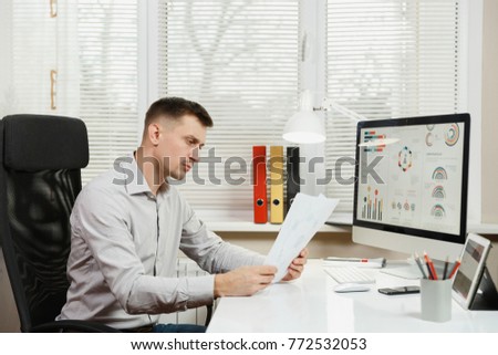 Serious and engrossed business man in shirt sitting at the desk, working at computer with modern monitor, folders, coffee or tea, documents in light office on window background. Manager or worker