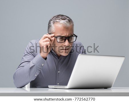 Mature businessman working with a laptop and having vision problems, he is staring closely at the computer screen and adjusting his glasses