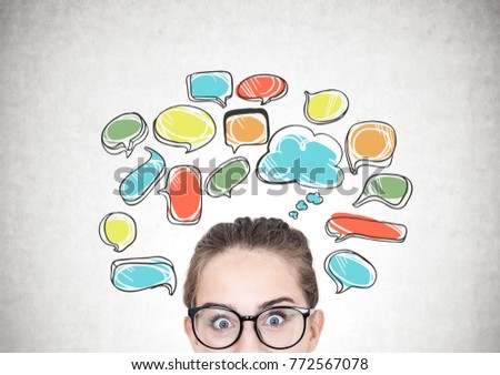 Close up of a teenage girl with fair hair wearing glasses and looking at the viewer in astonishment. Concrete wall background with colorful speech bubbles. Mock up