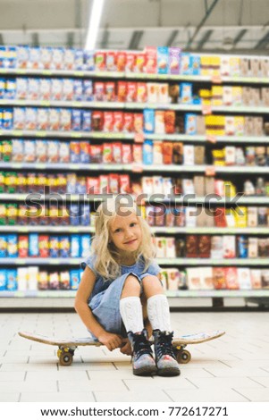 adorable blonde kid sitting on skateboard in supermarket with shelves behind