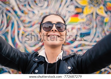 Smiling young woman taking a selfie with the Berlin Wall in the background.