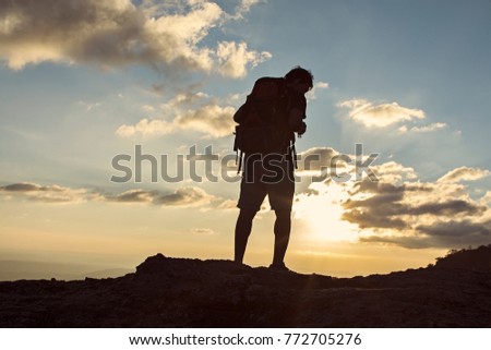 Man with backpack standing on cliff at sunset time, sun rays on background with clouds, this man looking at view,  warm tone and almost in silhouette photo