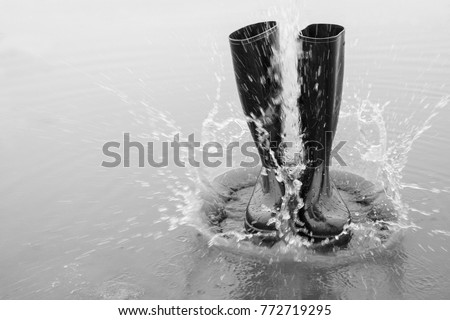 Black rubber boots fall into a puddle and the water splashes. Wellies are fashionable and the right footwear for bad weather. A black and white picture. Rubber boots become art.