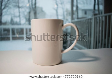 A tall white coffee mug in front of snow scene window on white surface.