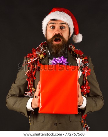 Business and celebration concept. Businessman with excited face holds present box and decorations. Man in smart suit, Santa hat and garlands on brown background. Manager with beard holds red gift.