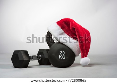 A large black Kettlebell wearing a red  Santa hat with a dumbbell next to it on a plain light gray background 