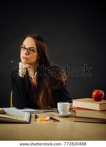 Photo of a teacher or business woman in her 30's sitting at a desk in front of a large blackboard.
