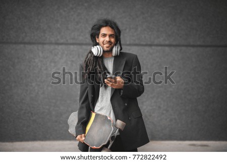 Close up of stylish handsome happy dreadlocks skater man in a suit standing with skate and mobile in hands and looking at the camera.