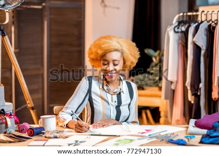 Young african fashion designer working with clothes drawings sitting at the office interior with different tailoring tools