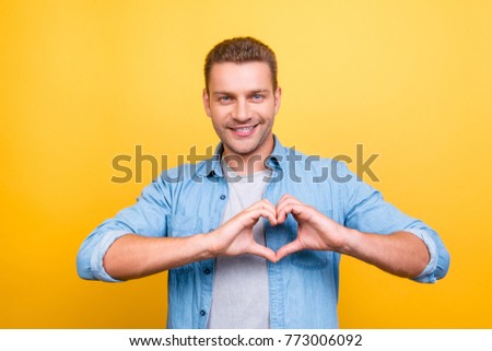 Portrait of  smiling, stunning man with stubble showing heart figure with fingers over yellow background