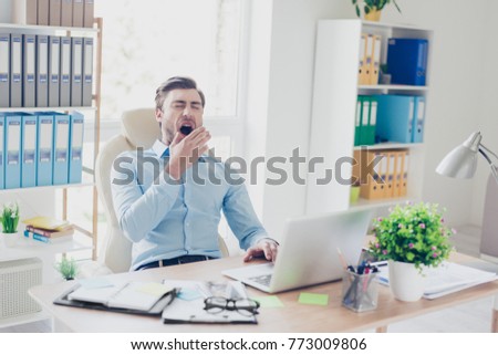 Portrait of sleepy tired agent wearing blue shirt, he is yawning and covering his mouth with a palm, he is sitting in front of computer