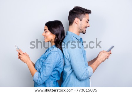 Beautiful, pretty, cute woman and handsome man in jeans shirts standing back to back and writing sms through 3g internet on smart phones over grey background Royalty-Free Stock Photo #773015095