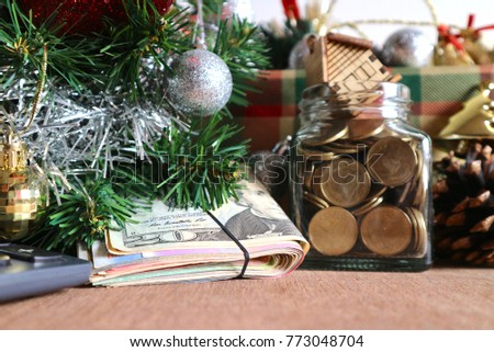 Wad of money under Christmas tree decorated with silver ribbons, glitter gold balls, calculator, money in glass bottle with miniature wooden house and gift box, dry pines on wood table               