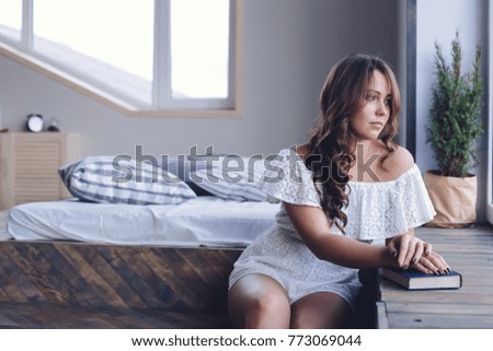 A girl reads a book, sitting in the morning next to a window and a bed