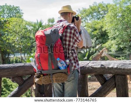 Young man photographer or traveler wearing hat and backpack with shoe and drinking water plastic bottle, holding camera and taking photo of giraffe in a sunny day. Hobby lifestyle concept.