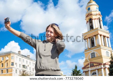 Young joyful woman taking self-portrait photograph with easily handled camera against bell tower of church in Russia