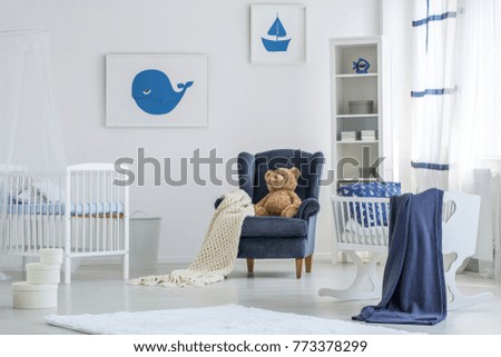 Blue blanket on cradle and teddy bear on armchair in white baby's bedroom with marine posters