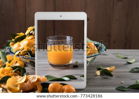 White tablet with glass of juice on screen standing in front of ceramic dish filled with tangerine peels on gray wooden table. Creative composition with space for text. Health food concept.
