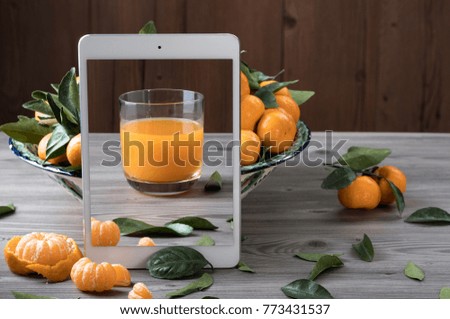 White tablet with glass of juice on screen standing in front of ceramic dish filled with fresh tangerines on gray wooden table. Creative photo composition. Health food concept. 