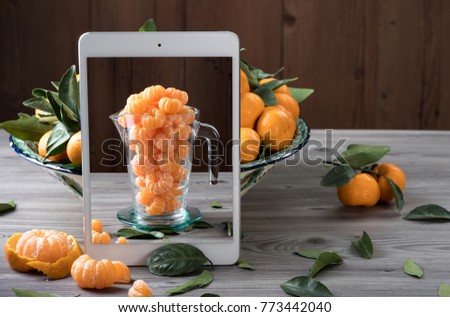 White tablet with peeled tangerines in glass jug on screen standing in front of ceramic dish filled with fresh tangerines on gray wooden table. Creative composition. Health food concept.