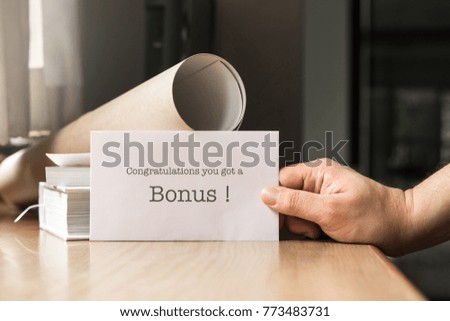 A man hand giving white envelope with message "Congratulation you got a bonus" on wooden working desk, which have two book and roll paperwork and soft light in morning