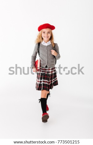 Full length portrait of a happy little schoolgirl dressed in uniform with backpack holding book while walking and looking at camera isolated over white background