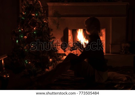 Dark photo in a dark room. A girl sitting by the burning fire of a fireplace. near the decorated Christmas tree with burning lights garlands. Home simple and cozy atmosphere.

