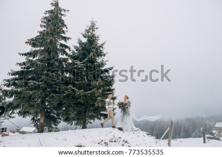 Beautiful couple stands between huge snowy pines, woman in grey wedding dress and long veil, man in sweater