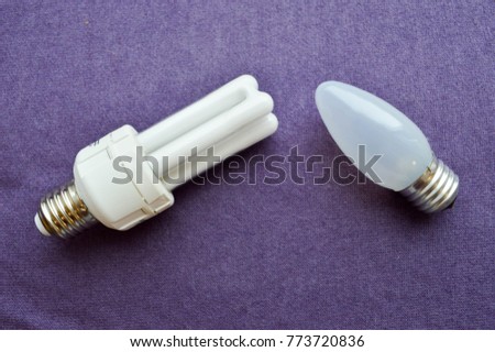 Two light bulbs. White energy-saving fluorescent bulb with four tubes, with a silver base and an oblong incandescent bulb with gray glass on a purple cloth background.