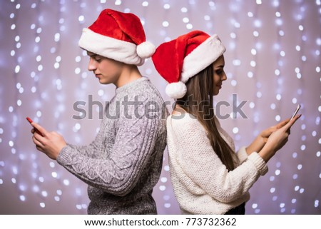 Young couple are browsing, typing, reading at their phones, standing back to back, wearing santa hat on light bokeh background