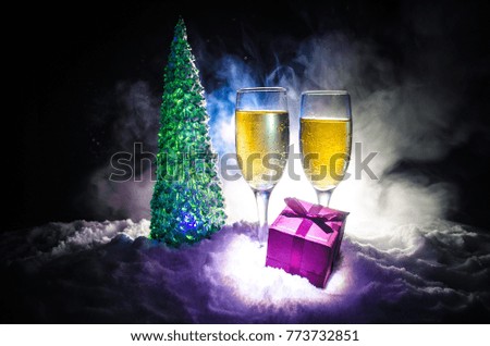 New Years Eve celebration background with pair of flutes and bottle of champagne with christmas tree on snow on dark background. Selective focus