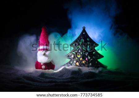 Happy Santa Claus Doll on Christmas time with tree and snow. Colorful bokeh background. Santa Clause and Merry Christmas model figure toy on dark toned foggy background. Selective focus