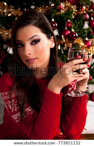 Beauty young girl with red wine glass in hand and red sweater and Christmas tree in background. Xmas and New Year Holiday concept