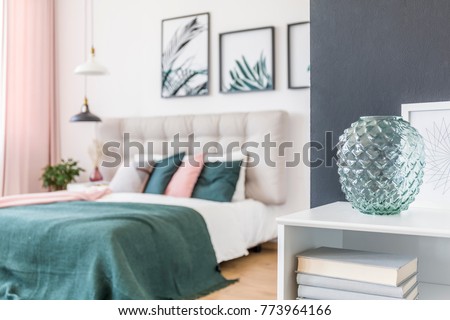Close-up of green glass vase on white shelf with books in green bedroom interior with lamps and gallery of posters
