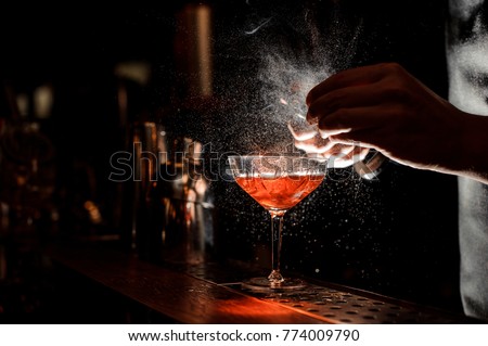 Barman`s hands sprinkling the juice into the cocktail glass filled with alcoholic drink on the dark background Royalty-Free Stock Photo #774009790